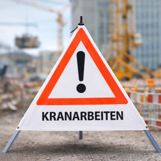 Faltsignal Warnschild mit rotem Dreieck und Ausrufezeichen, Text KRANARBEITEN, Baustelle im Hintergrund