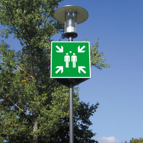 Grünes Sammelstellenschild mit zwei Personen und vier Pfeilen nach innen an Laternenpfahl vor blauem Himmel und Bäumen