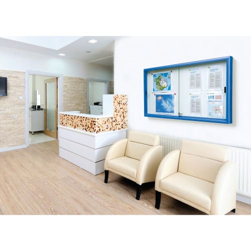 Schaukasten KLASSIK mit blauer Rahmen und Schiebetüren an weißer Wand in modernem Wartebereich mit zwei cremefarbenen Sesseln