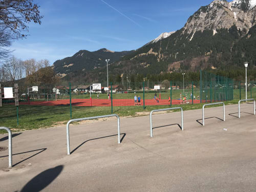 Anlehn- und Absperrbügel aus Stahl zum Einbetonieren mit Ø 60 mm, auf einem Gehweg vor einem Sportplatz mit Bergen im Hintergrund