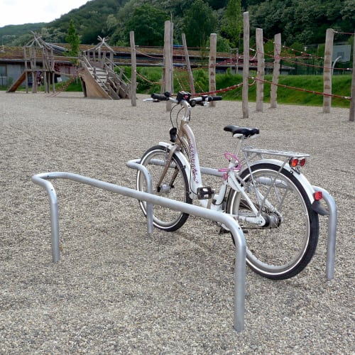 Anlehnbügelparker für Fahrräder mit einem silbernen Fahrrad auf einem Kiesplatz vor einem Spielplatz