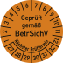 Orange Prüfplakette mit der Aufschrift Geprüft gemäß BetrSichV und Nächster Prüftermin, mit Zahlen von 1 bis 12 und 27 bis 32
