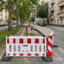 Klemmbake für Schrankenzaun EURO 2 Absperrung auf einer Baustelle in einer Stadtstraße mit Bauarbeitern und Baumreihe
