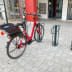Fahrradständer Lambda® aus Metall mit einem roten Fahrrad davor auf gepflastertem Boden vor einem Gebäude