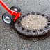 NESTLE Kanaldeckelheber mit roten Griffen und schwarzen Rädern hebt einen runden Kanaldeckel aus Asphalt
